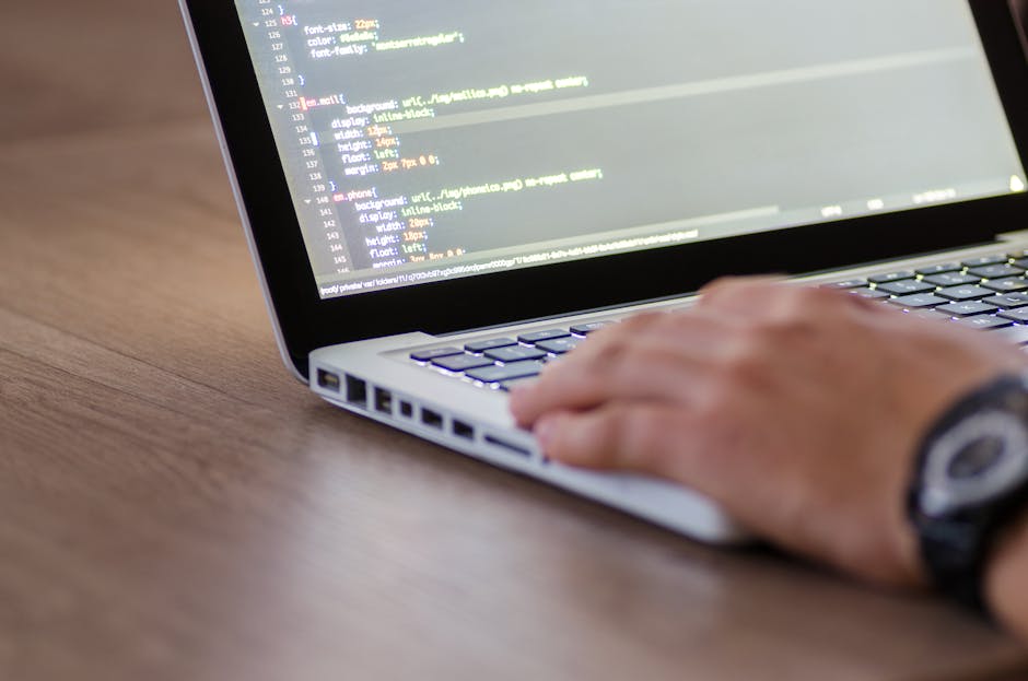 A person with a wristwatch is using a laptop placed on a wooden surface, displaying a screen with lines of colorful code and script in a text editor. The keyboard is illuminated with backlit keys, and